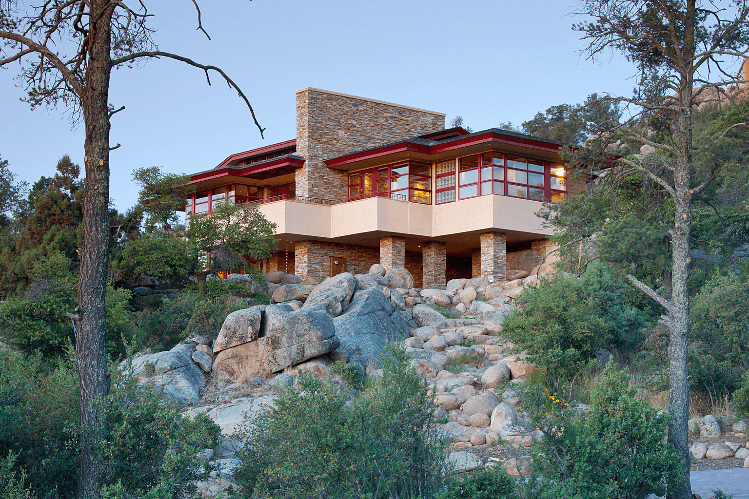 A multi-level stone-and-stucco house with red window frames, lit at dusk atop a hillside of granite boulders and pines.