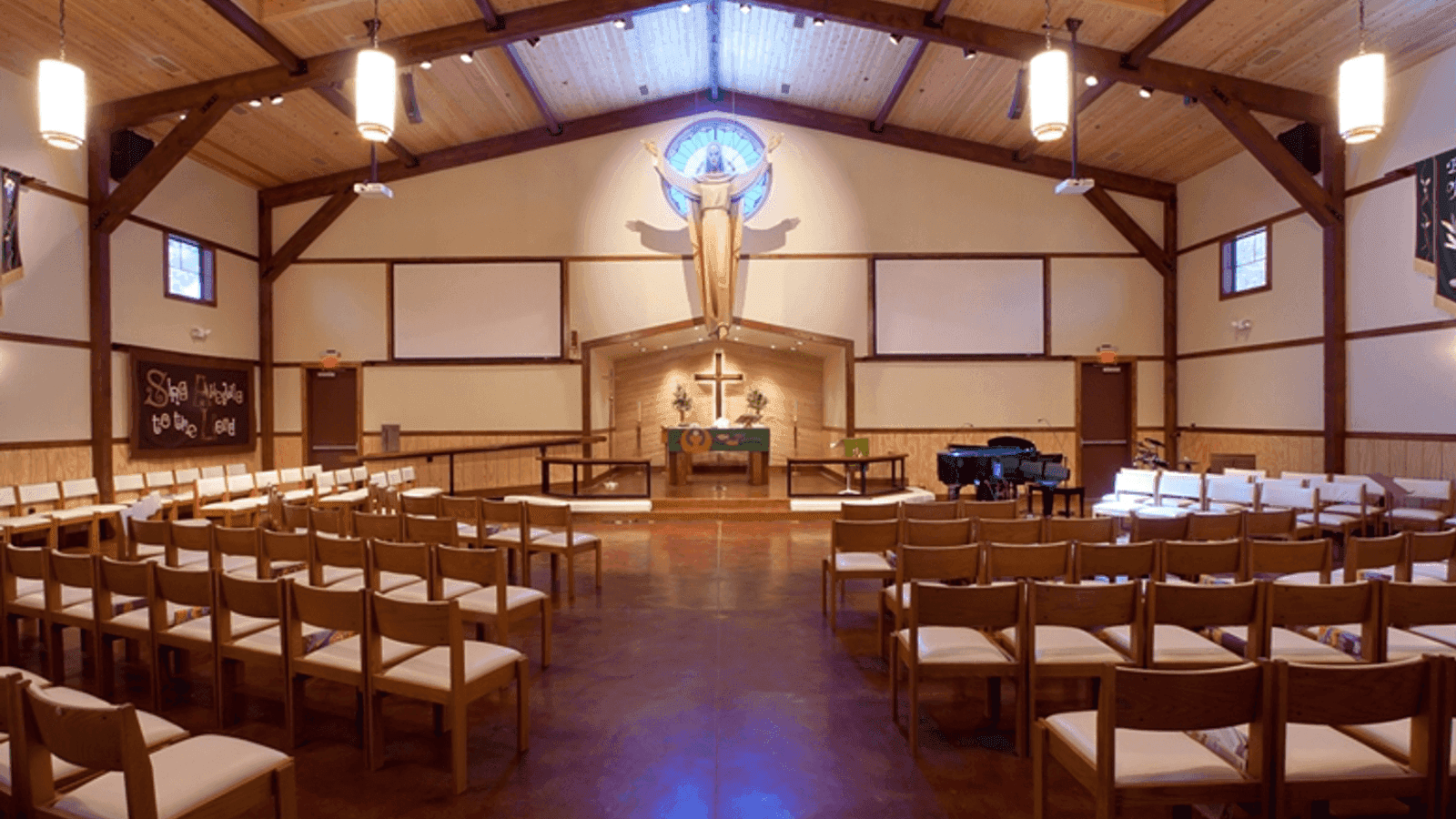 The interior of a chapel with exposed timber trusses, rows of pale chairs, and a stained-glass window above the altar.
